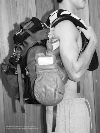 Athletic man with bare torso and backpack. Gloves and towel hanging. Gray sweatpants and wood background. Black and white.