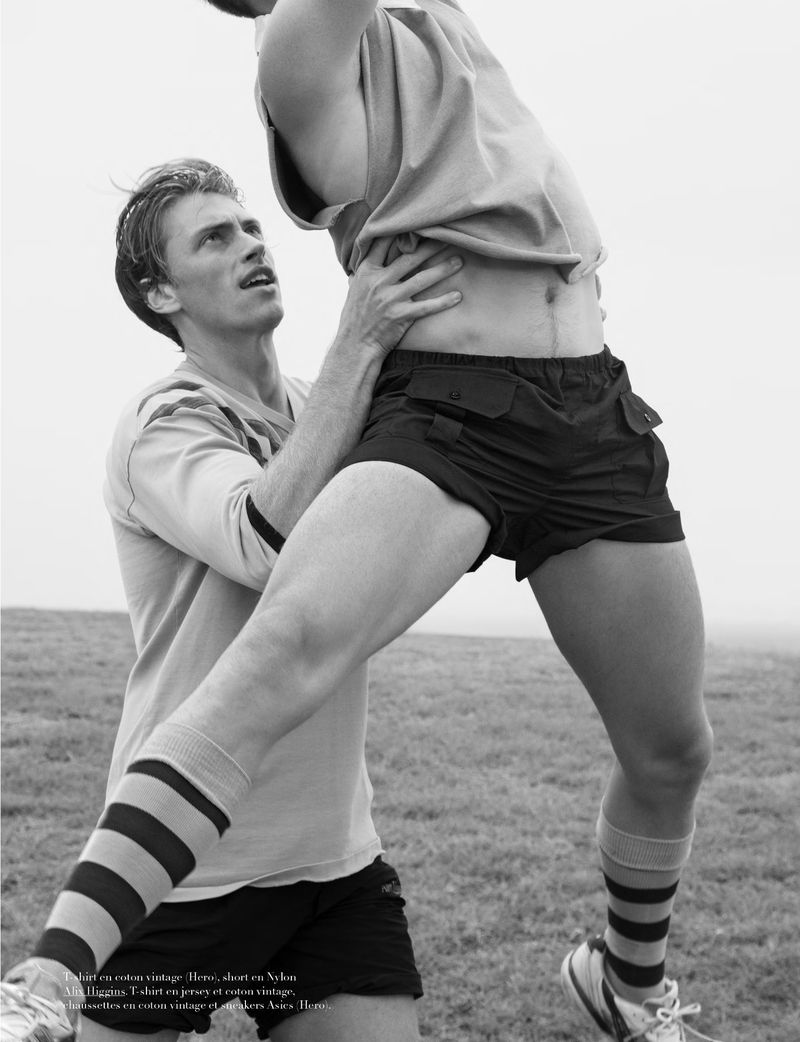 Monochrome shot of two men in athletic wear on a field; one man is lifting the other up, with a bright sky overhead.