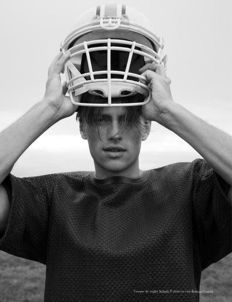 Portrait of a young man wearing a dark textured shirt and holding an American football helmet above his head in an outdoor setting.
