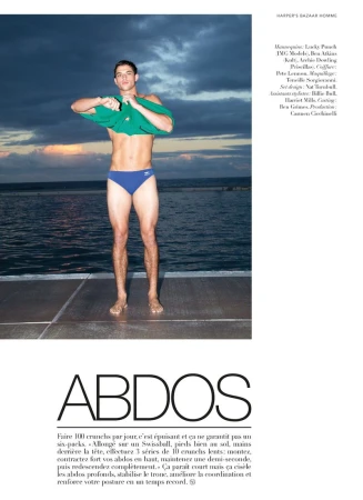 Man standing on a dock by the water at sunset removing a green t-shirt while wearing blue swim briefs for Harper's Bazaar France Homme