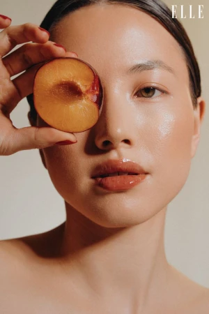 Close-up portrait of a woman holding a plum slice to her eye, showcasing glossy skin, and nude lipstick, with magazine logo 'ELLE' in the corner.