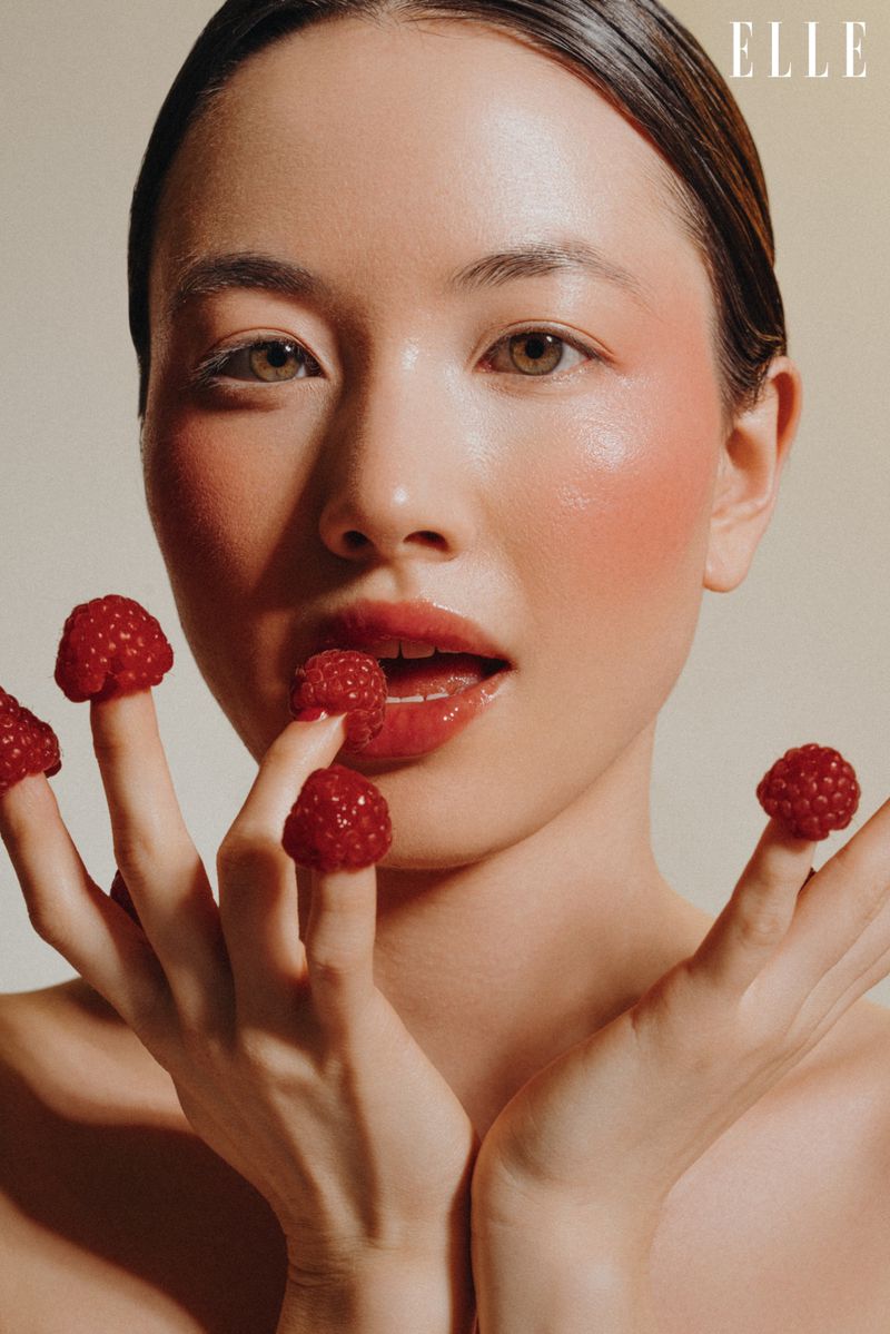Glamorous portrait of an Asian woman with raspberries on her fingers and glossy lips, against a neutral background. Magazine 'Elle' logo.