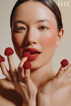 Glamorous portrait of an Asian woman with raspberries on her fingers and glossy lips, against a neutral background. Magazine 'Elle' logo.