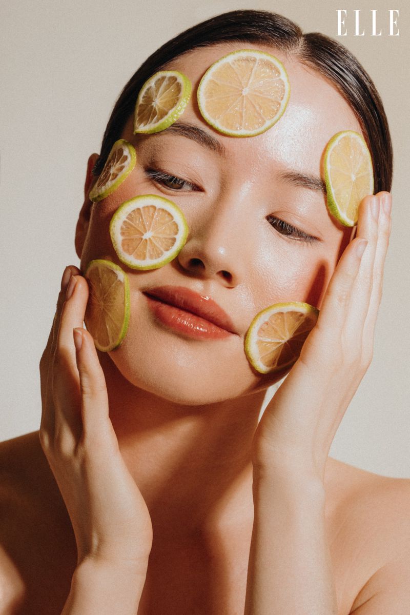 Asian woman with lemon slices on her face in a beauty shot with glossy lips, resting her hands on her face in a studio setting.