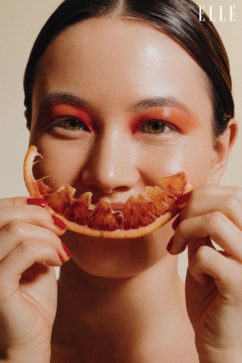 Close-up of woman holding orange slice over mouth with bright orange eye makeup, red nails, and copy space. Beauty, Fashion, ELLE.