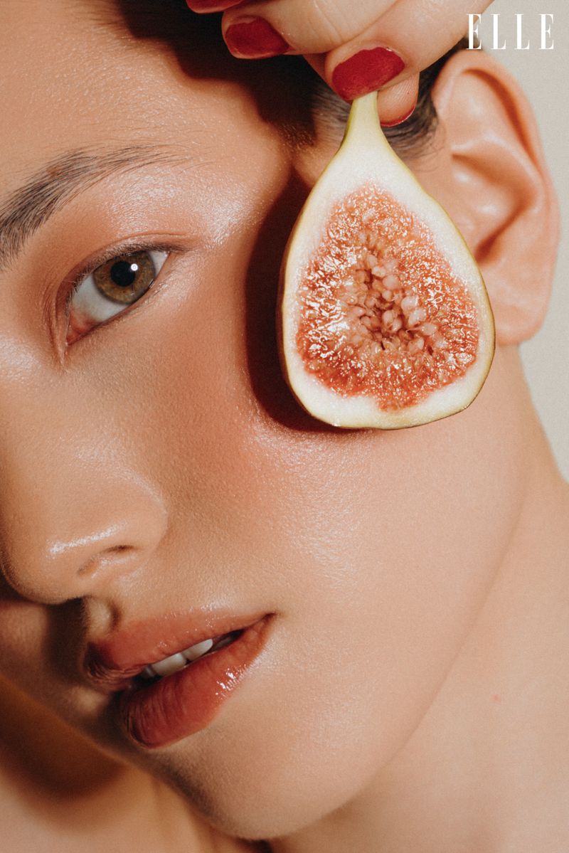 Close-up studio shot of a light-skinned adult female's face partially obscured by a halved fig held by a hand with red nail polish.