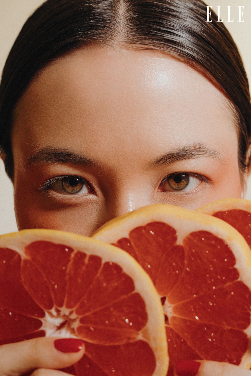 Close-up fashion shot of a young woman with red nail polish holding grapefruit slices, against a neutral background, featuring the Elle logo.