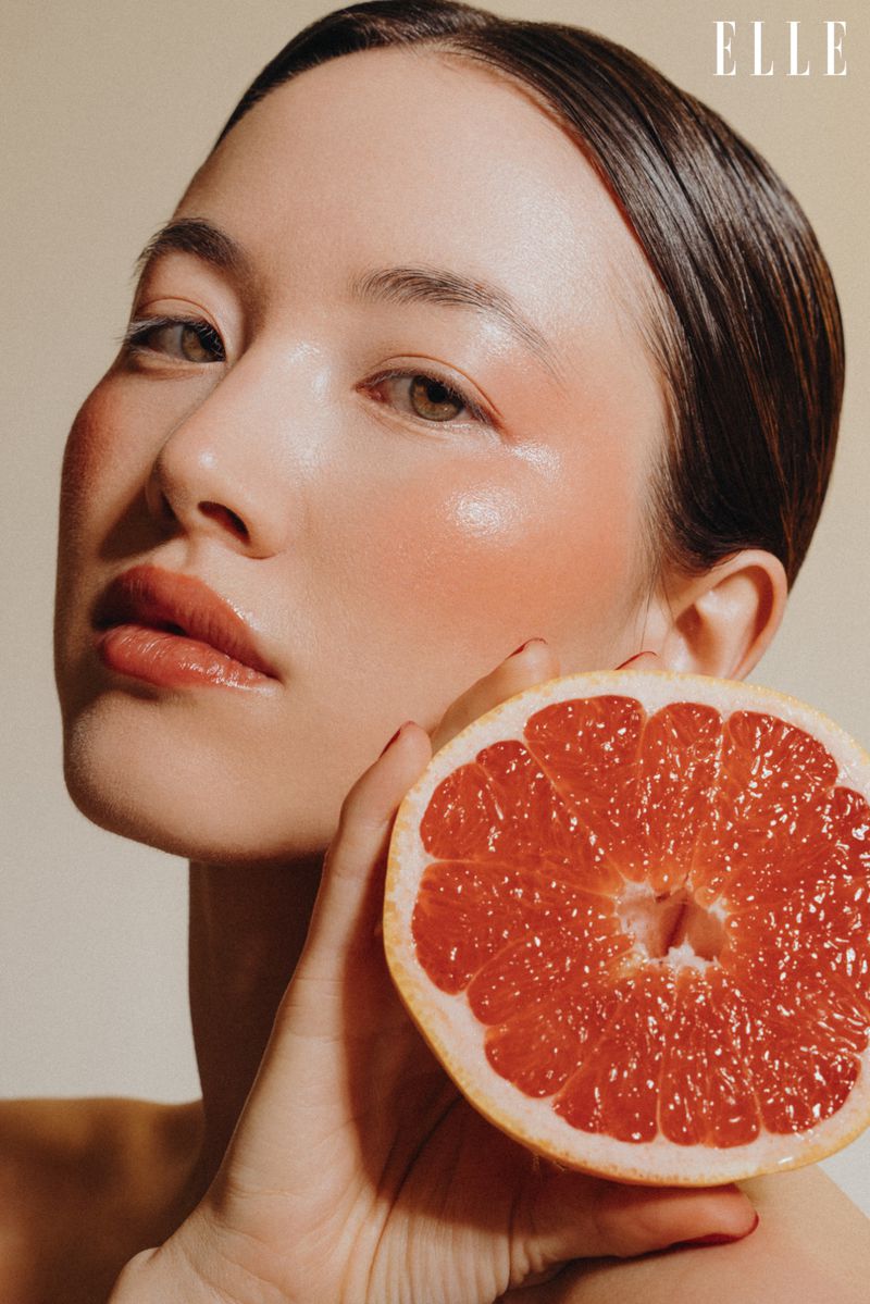 Close-up of a woman with blush, holding a halved grapefruit next to her face, against a neutral background, and the ELLE logo at the top.