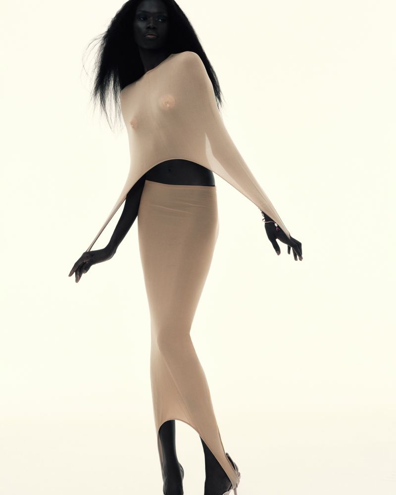 A Black female fashion model poses in a light tan, sheer, fitted dress against a white backdrop in a studio shot with minimalist lighting.