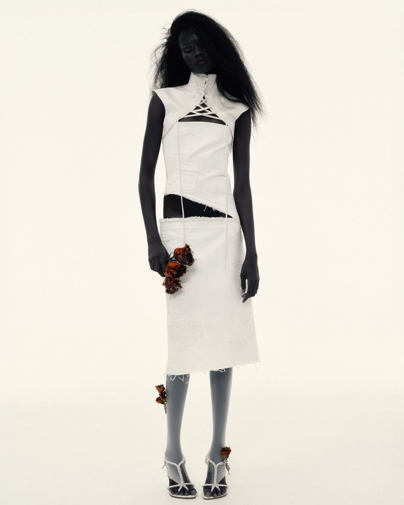 A dark-skinned fashion model in a cut-out white dress poses against a white backdrop while holding wilted roses. Studio shot.