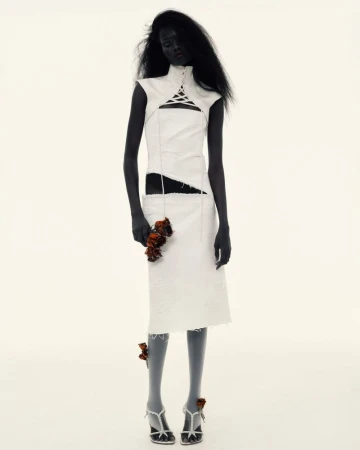 A dark-skinned fashion model in a cut-out white dress poses against a white backdrop while holding wilted roses. Studio shot.