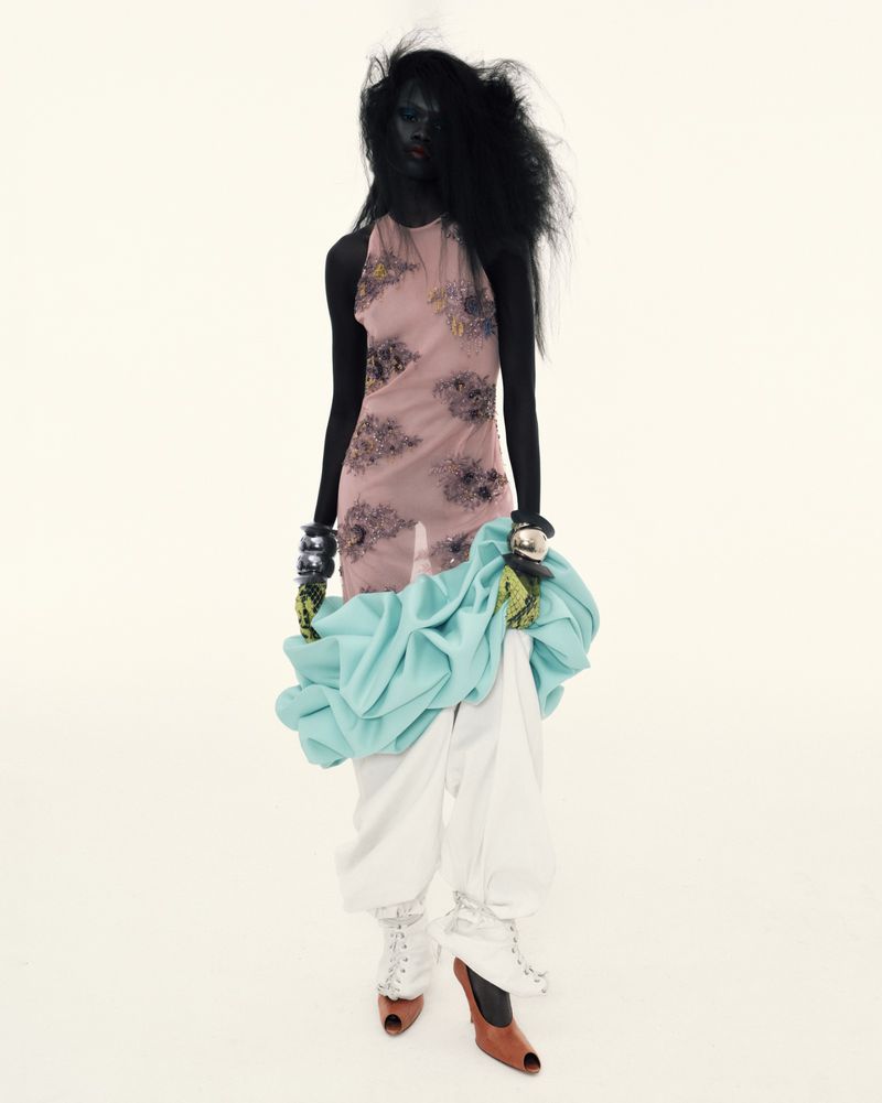 A dark-skinned fashion model poses in a halter top and white pants against a bright white backdrop in a studio fashion editorial.