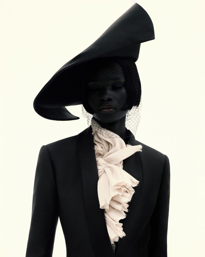 A person wearing a black suit, wide-brimmed hat with netting, and ruffled collar stands against a stark white backdrop in a studio portrait.