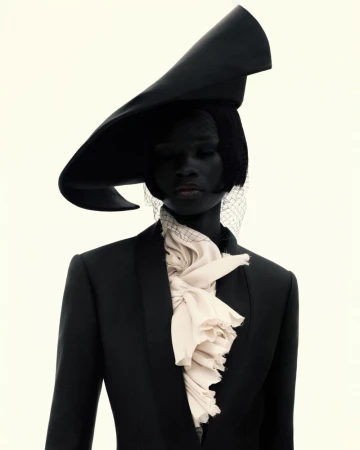 A person wearing a black suit, wide-brimmed hat with netting, and ruffled collar stands against a stark white backdrop in a studio portrait.