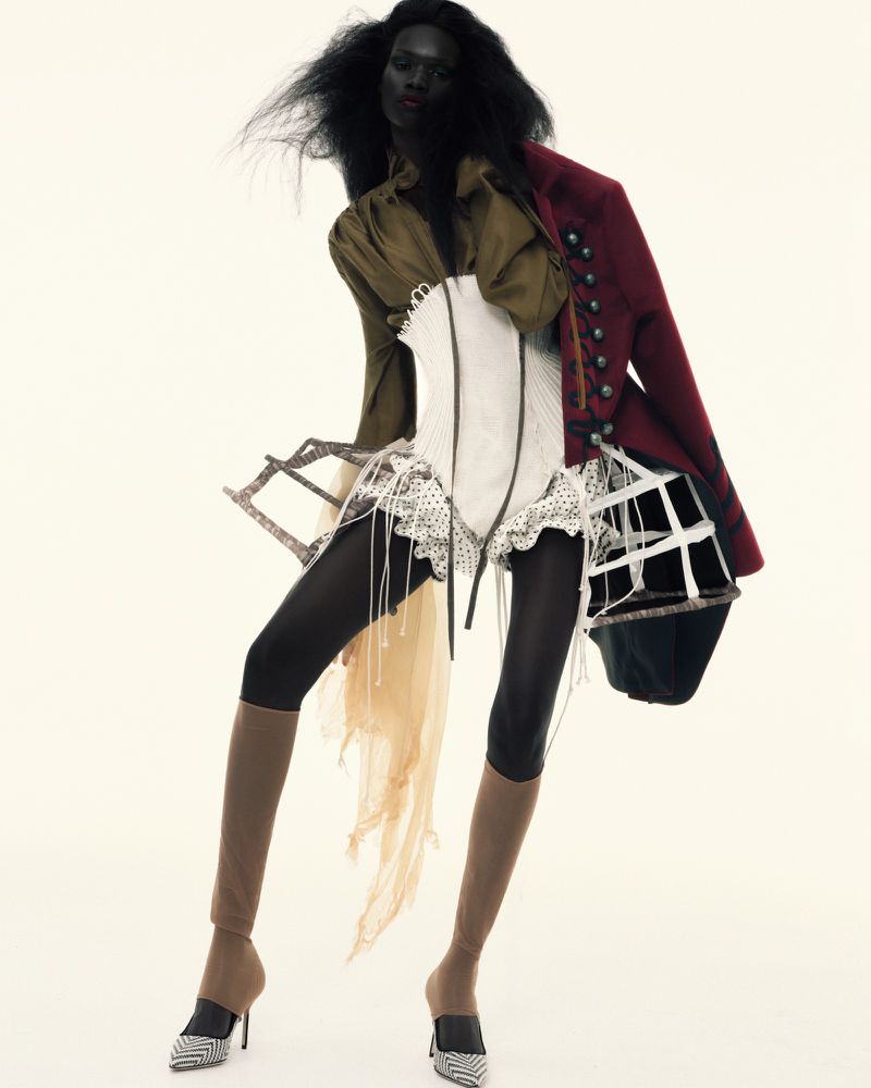 An adult female fashion model with dark skin and voluminous dark hair poses in a studio wearing an eclectic mix of garments and heels.