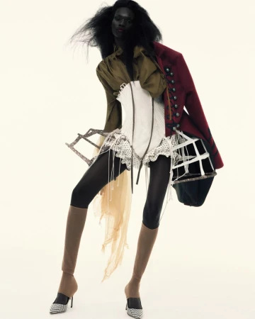 An adult female fashion model with dark skin and voluminous dark hair poses in a studio wearing an eclectic mix of garments and heels.