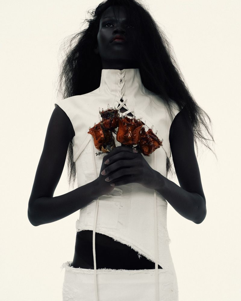 A woman with dark skin and hair poses with a bouquet of orange-brown flowers against a solid white background, wearing a white top and skirt.