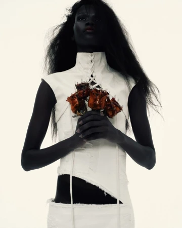 A woman with dark skin and hair poses with a bouquet of orange-brown flowers against a solid white background, wearing a white top and skirt.