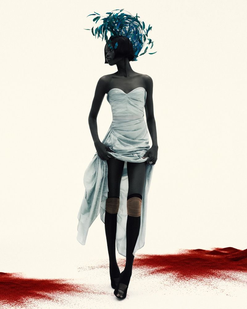 Fashion photo of an African American woman in a strapless dress and tall feathered headpiece, standing on a red dust surface.