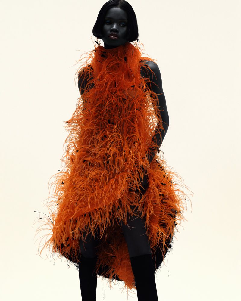 A single female fashion model poses in a vibrant orange feathered dress against a light background, showcasing high fashion style.