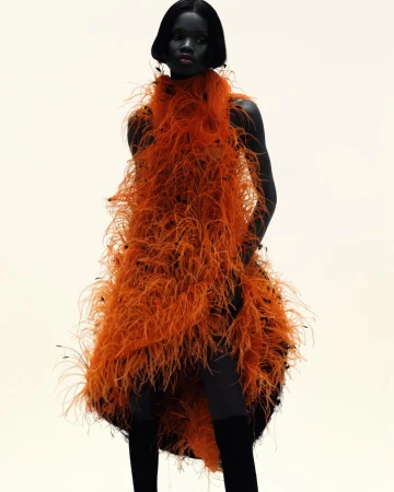A single female fashion model poses in a vibrant orange feathered dress against a light background, showcasing high fashion style.