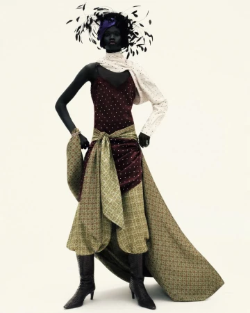 A woman in an ornate maroon and green dress with a feathered hat poses against a white background in a fashion editorial shot.