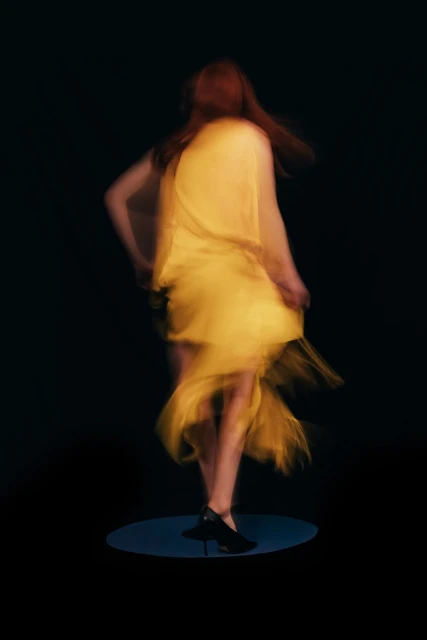 Back view of a woman with red hair wearing a yellow dress and black high heels, captured in a long exposure against a black backdrop.