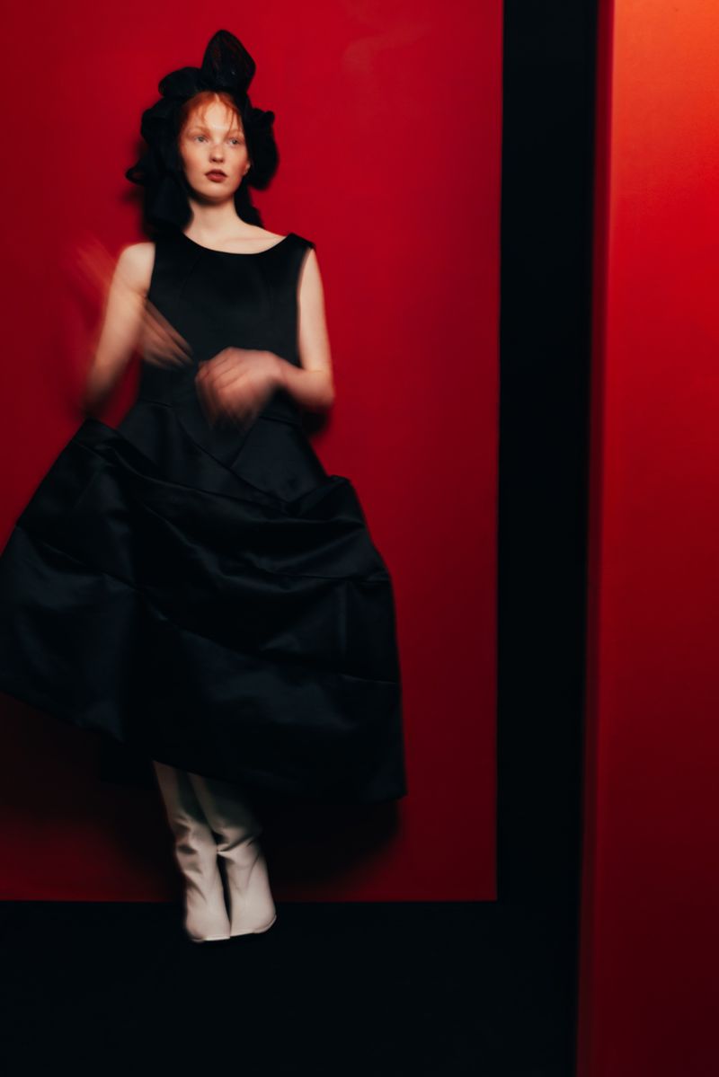 A woman in a black dress poses against a vibrant red backdrop, exuding high fashion and style, complemented by white boots and dramatic lighting.