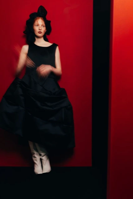 A woman in a black dress poses against a vibrant red backdrop, exuding high fashion and style, complemented by white boots and dramatic lighting.