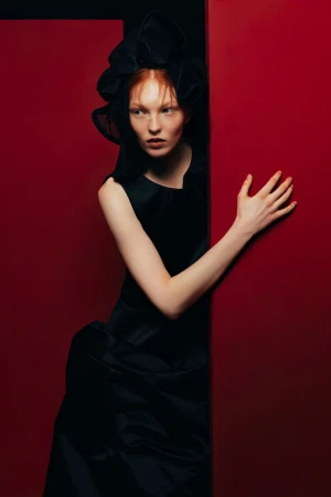 Elegant redhead woman poses in a black dress and elaborate headwear against a red background in a studio shot for a fashion editorial.