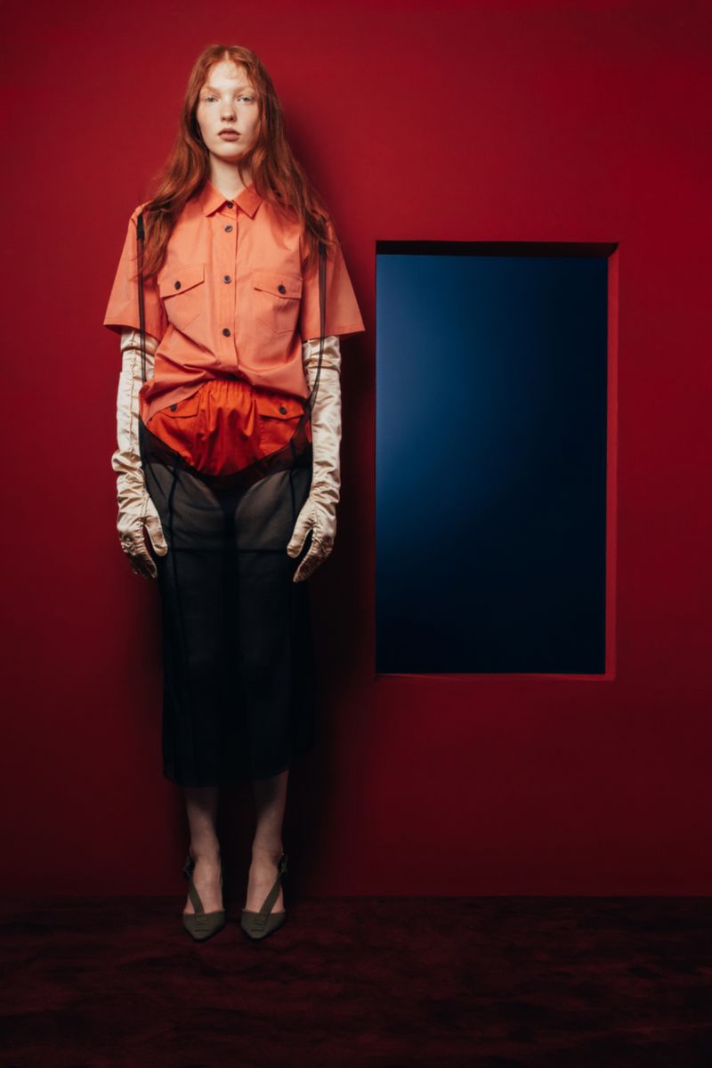 A red-haired model poses in a pink shirt and black skirt in front of a red wall with a blue square, in a studio setting.