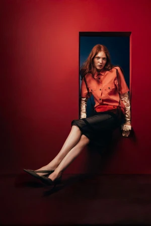 Fashion photo features a red-haired woman in an orange button-down shirt and black skirt, seated in a doorway against a red wall.