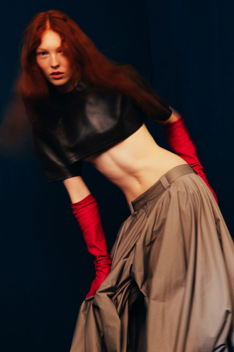 Fashion shot of a red-haired woman with bare stomach, wearing a black crop top, flowing grey skirt, and red gloves, posing indoors.