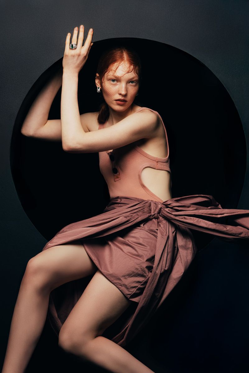 Red-haired woman in stylish pose, wearing a pink bodysuit and draped dusty rose skirt in a fashion studio setting, high angle.