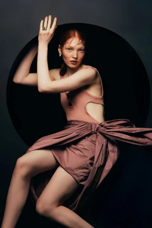 Red-haired woman in stylish pose, wearing a pink bodysuit and draped dusty rose skirt in a fashion studio setting, high angle.