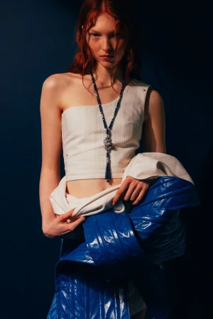 Young woman with red hair in a one-shoulder top and shiny blue skirt poses against a dark blue backdrop in a fashion-style portrait.