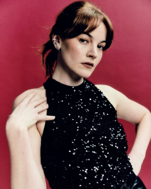 Portrait of a young woman with dark brown hair, wearing a black sequined top and silver earrings, posing against a red background.