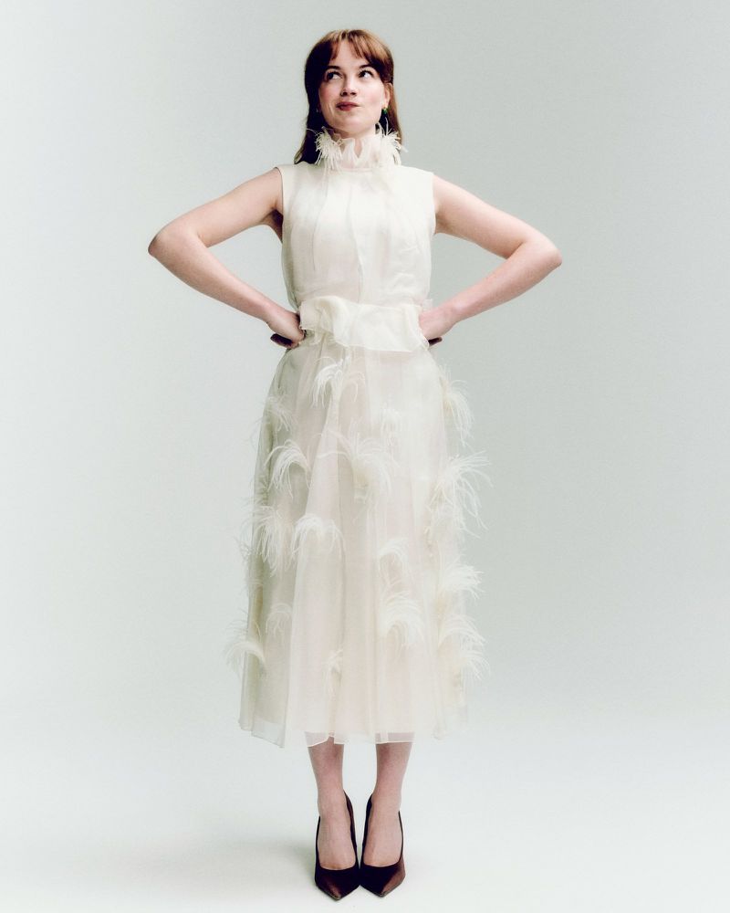 A woman in a sleeveless formal dress with feathers stands in a photo studio, hands on hips, looking up and to the side with a slight smile.