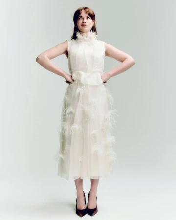 A woman in a sleeveless formal dress with feathers stands in a photo studio, hands on hips, looking up and to the side with a slight smile.