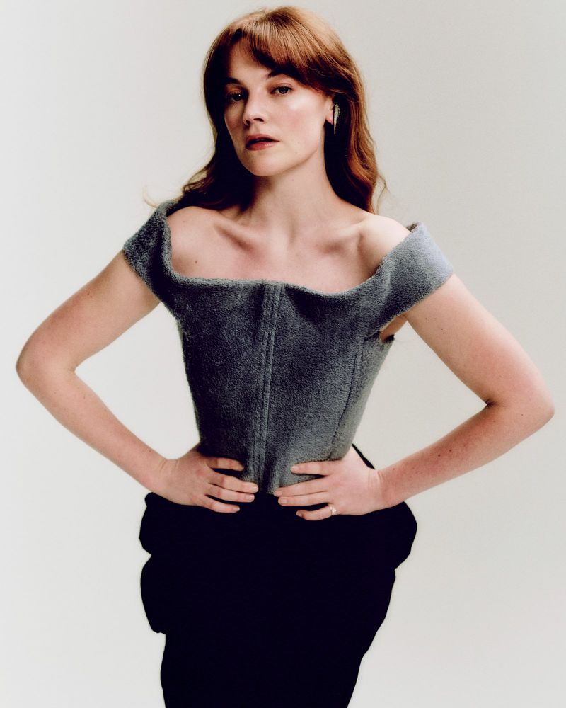 Portrait of a red-haired woman in a gray corset and black skirt, looking at the camera with a neutral expression against a white backdrop.