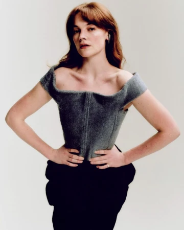 Portrait of a red-haired woman in a gray corset and black skirt, looking at the camera with a neutral expression against a white backdrop.