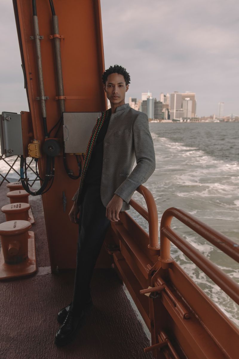 Fashionable adult poses on a boat with city skyline background wearing a grey jacket, black shirt and pants for outdoor photoshoot.
