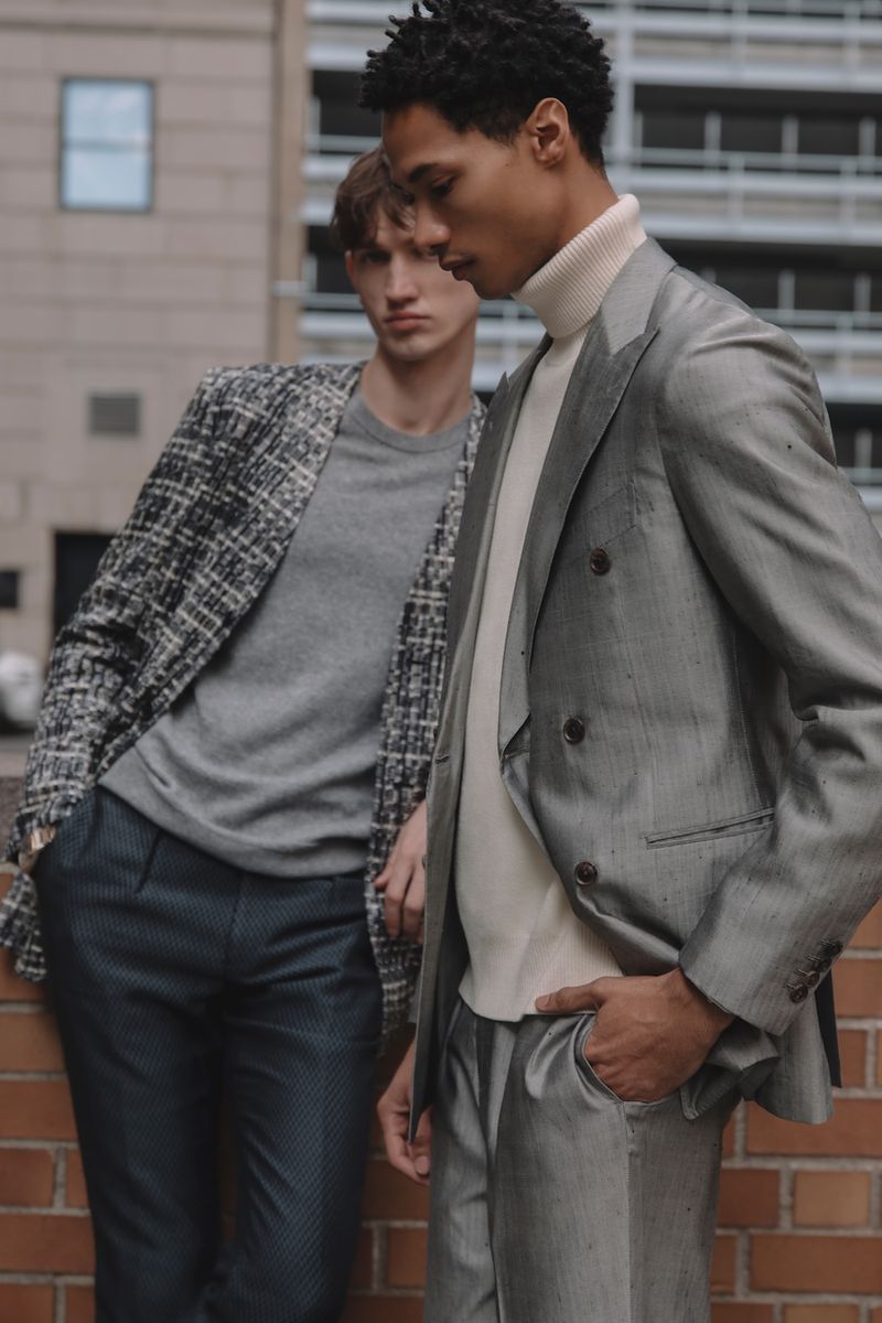 Two men in stylish blazers and trousers stand confidently against a brick wall and building. Fashion editorial shot, urban scene.
