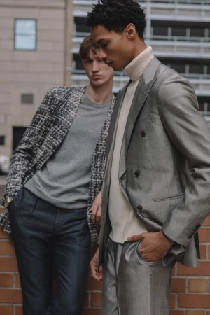 Two men in stylish blazers and trousers stand confidently against a brick wall and building. Fashion editorial shot, urban scene.