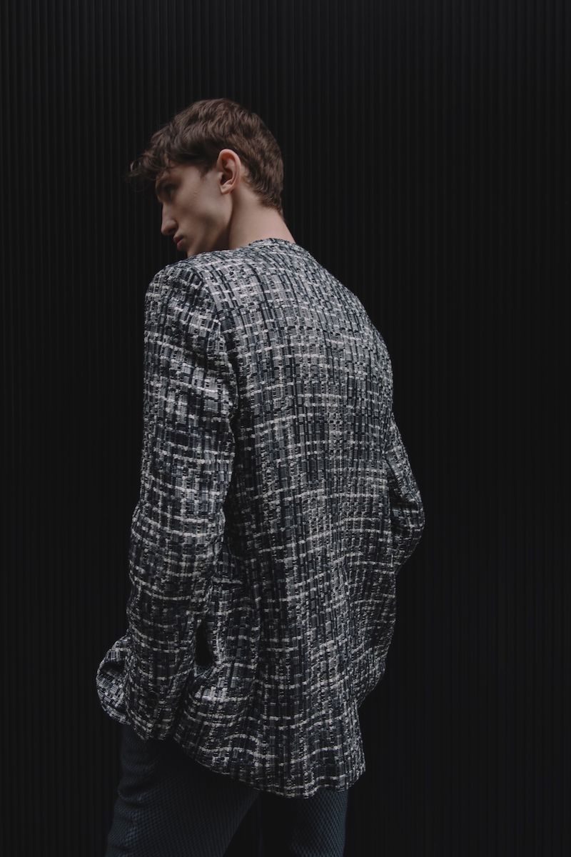 Man in tweed jacket against a black background. The jacket is black and white. The man is turned away from the viewer but looking back.