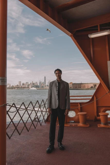 Man in grey jacket and black pants stands on a ferry with a city skyline and water backdrop on a sunny day with orange ferry structure.