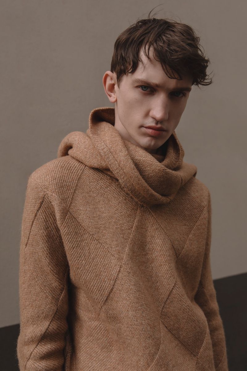 Portrait of a young adult man wearing a brown knit hoodie with geometric design, posing against a gray background in a fashion setting.