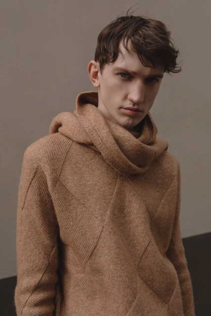 Portrait of a young adult man wearing a brown knit hoodie with geometric design, posing against a gray background in a fashion setting.