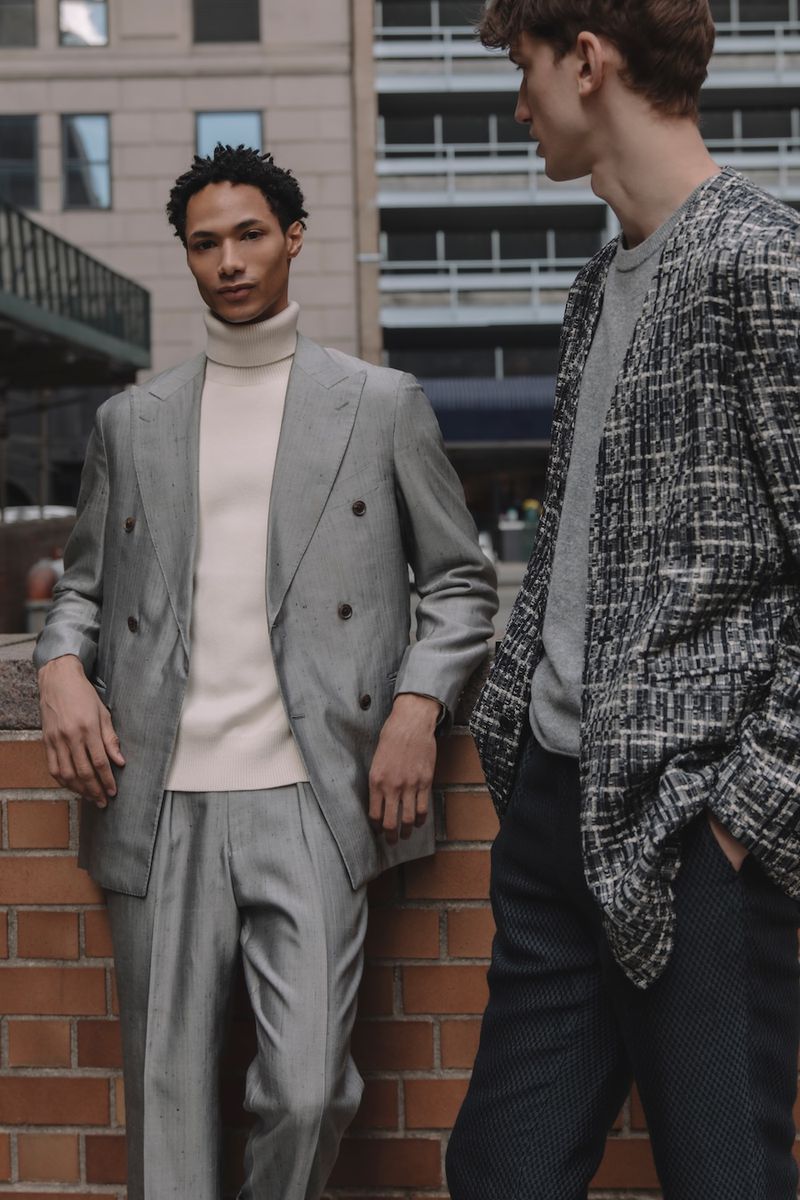 Two male models in tailored suits standing outside against a brick wall and city background for Kaltblut Magazine.