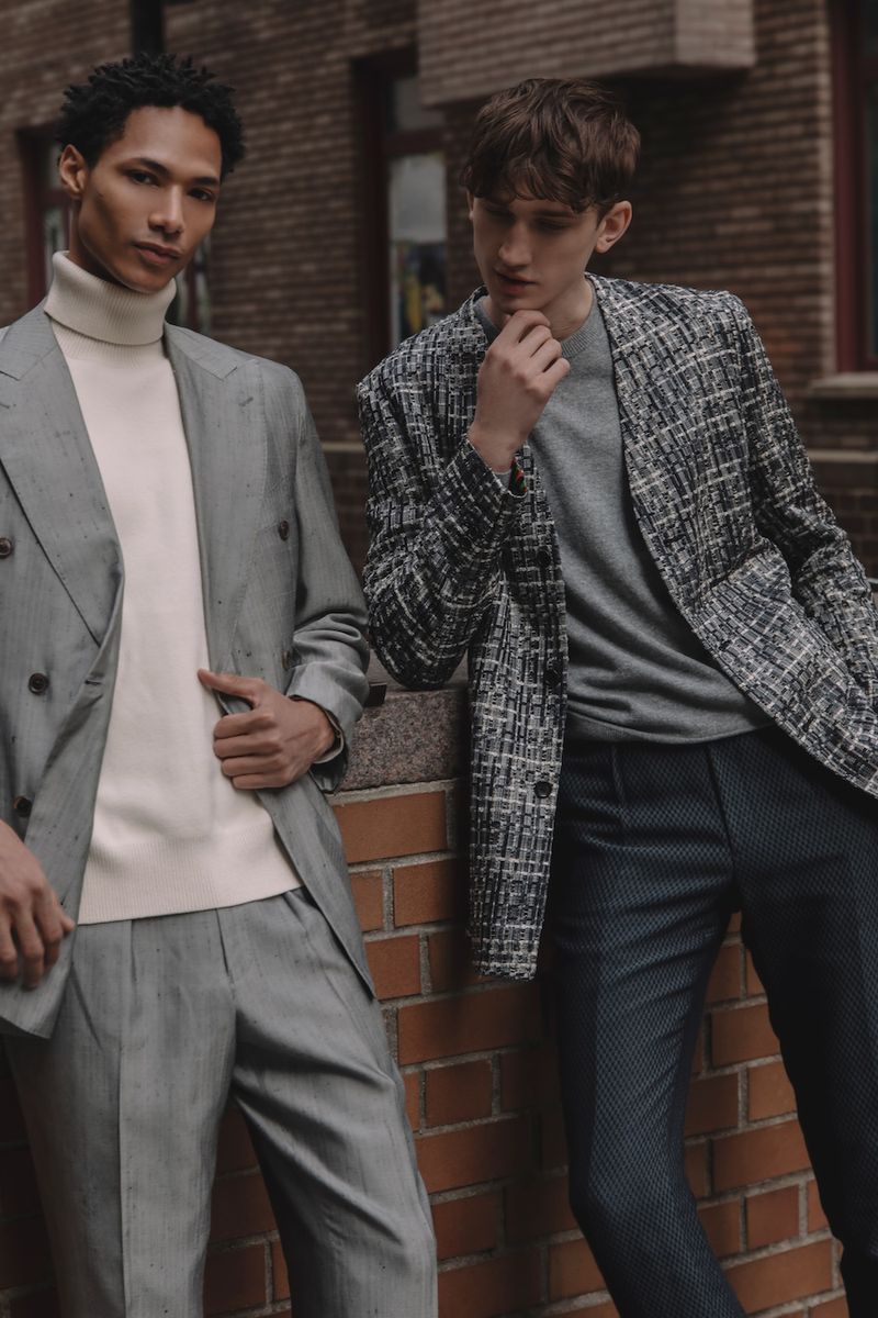 Two adult men in stylish blazers and dress pants pose against a brick wall in an urban setting, showcasing contemporary fashion.